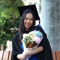 A young woman in graduation cap and gown sits on a bench seat in UQ's Great Court, clutching a bouquet of flowers with a small teddy bear inside it.